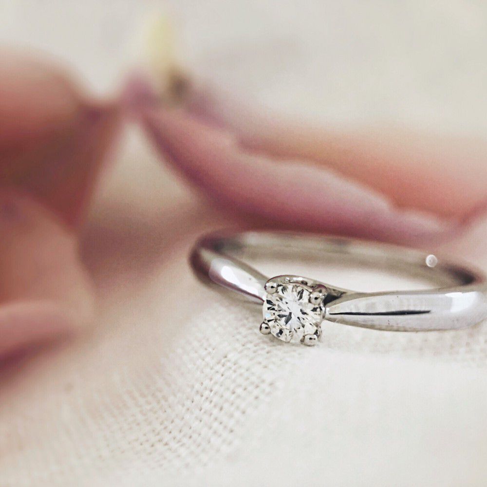 Een close-up van de Elegante 14 Karaat Witgouden Ring met Diamant van Excellent Jewelry, rustend op zacht licht stof met vervaagde roze bloemblaadjes op de achtergrond.