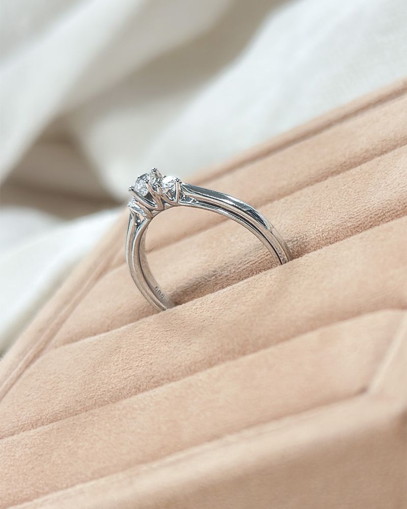 Een close-up van de Excellent Jewelry 14 karaat witgouden trilogie ring met 0,19 ct diamant, met een centrale en twee kleinere diamanten, rechtopstaand in een zachtbeige ringendoos tegen een stoffen achtergrond.
