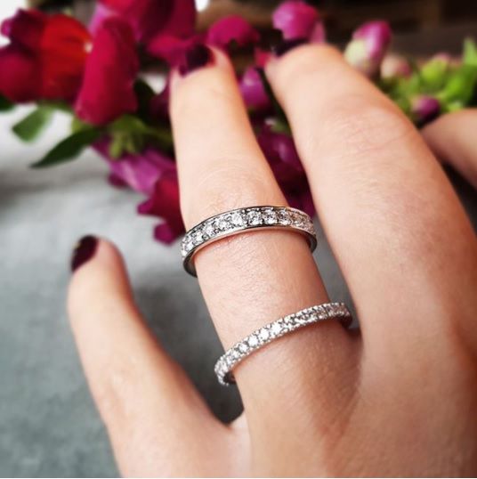 Een hand met donkerrode nagels draagt de Excellent Jewelry Witgouden Ring met Diamant: Tijdloze Verfijning in 14 Karaat en een andere diamanten ring aan de middelvinger, afgezet tegen vage roze bloemen.
