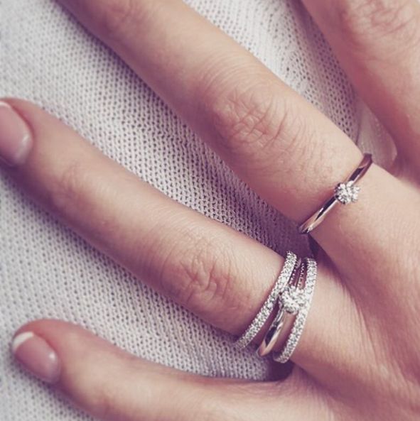 Een close-up van een hand die drie ringen draagt, waaronder de "Witgouden Ring met Diamant: Tijdloze Verfijning in 14 Karaat" van Excellent Jewelry, aan twee vingers tegen een lichte stoffen achtergrond.