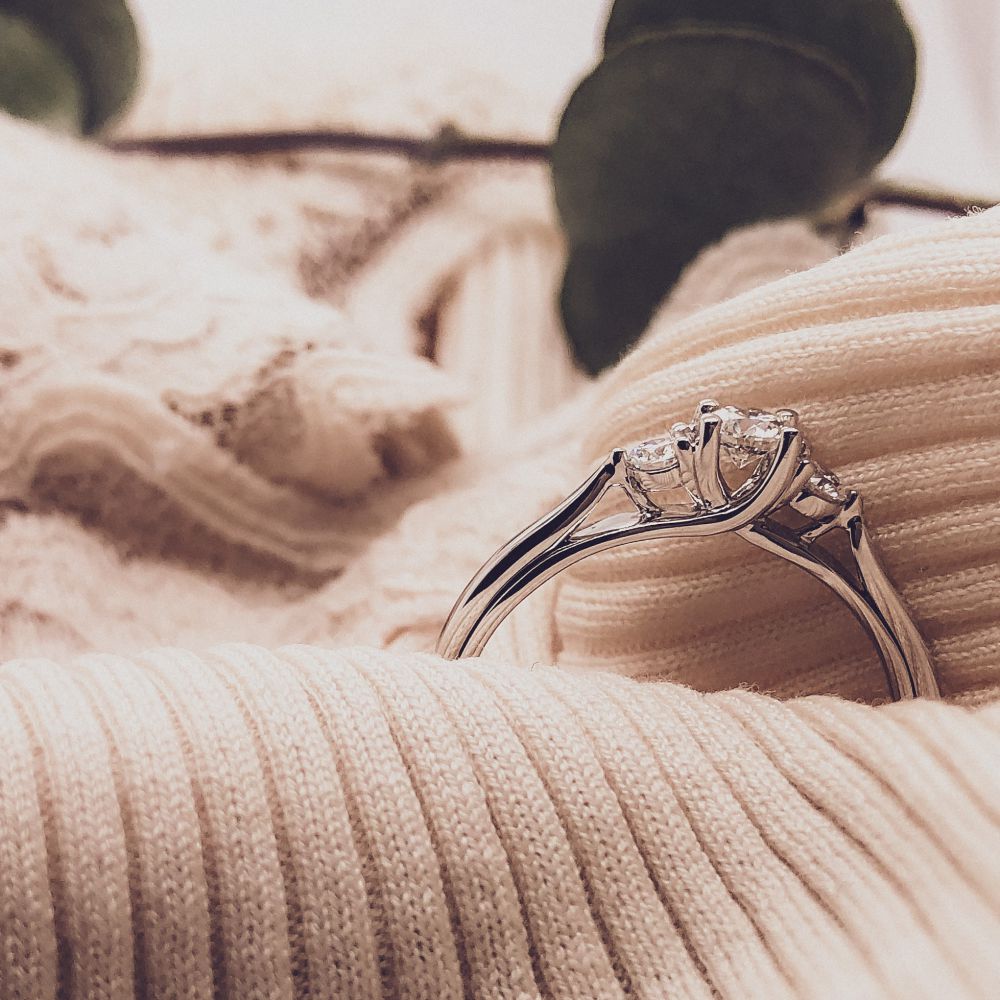 Een close-up van de Stijlvolle Witgouden Ring met Briljant Middensteen 0.31 crt. van Excellent Jewelry, liggend op zachte crème-kleurige stof met kantdetails en subtiel vervaagde groene bladeren op de achtergrond.
