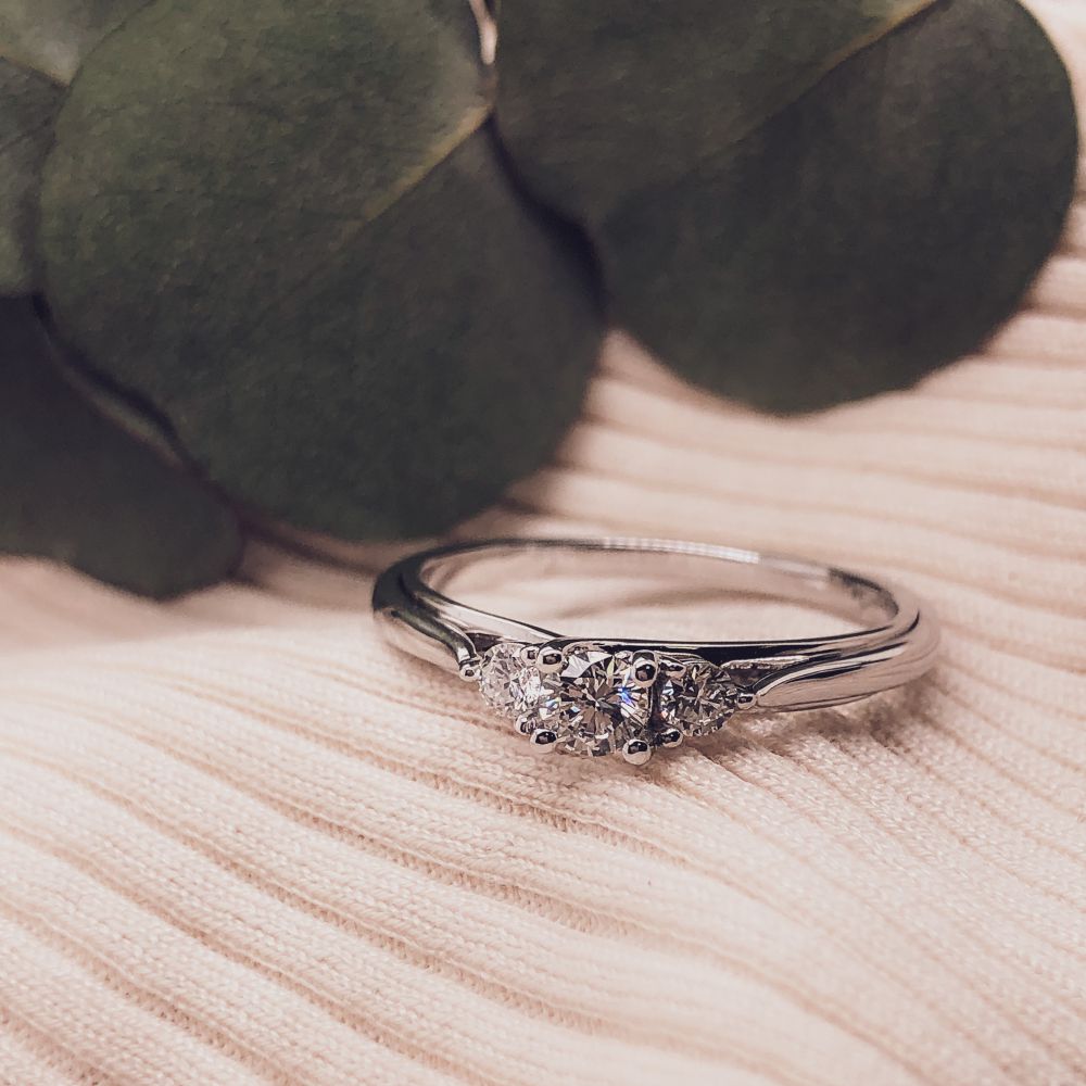 De Stijlvolle Witgouden Ring met Briljant Middensteen 0.31 crt. van Excellent Jewelry ligt op een bleek, geribd stoffen oppervlak, terwijl groene bladeren zacht vervaagd op de achtergrond verschijnen.