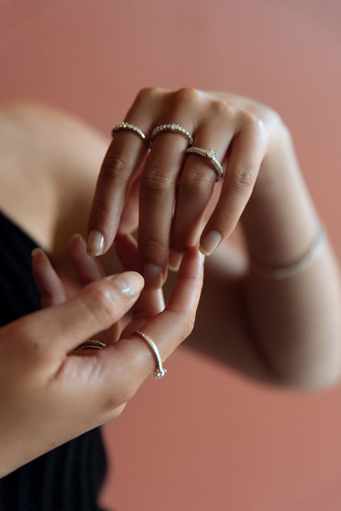 Een close-up van handen versierd met verschillende delicate ringen, waaronder de Elegante Witgouden Ring met Diamant van 0,32 crt van Excellent Jewelry, tegen een zachtroze achtergrond terwijl de ene hand de andere zachtjes aanraakt.