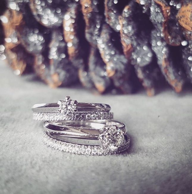 Twee Elegante Witgouden Ringen met Briljant (0,35 crt) van Excellent Jewelry rusten op een zacht grijs oppervlak, terwijl een onscherpe, fonkelende dennenappel op de achtergrond bijdraagt aan de zachte en romantische elegantie van de afbeelding.