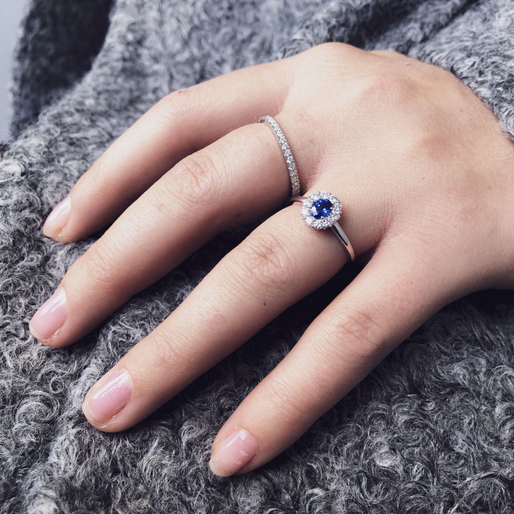 Een hand die de Excellent Jewelry Stunning White Gold Eternity Ring met 0,50 karaat diamanten en een zilveren ring met een blauwe edelsteen omringd door diamanten draagt, rust op een zachte, grijze stof met textuur.