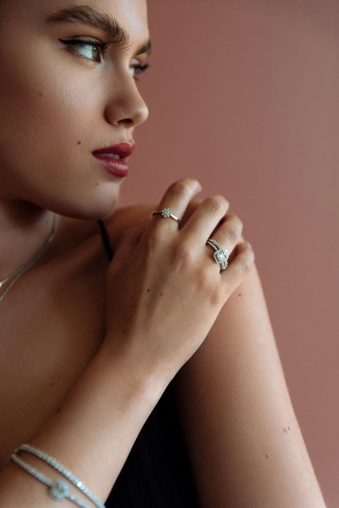 Een vrouw met opvallende wenkbrauwen en rode lippenstift kijkt opzij, draagt zilveren armbanden, een halsketting en de Elegant White Gold Ring with Brilliant and Baguette Diamonds 0.54 crt. by Excellent Jewelry, tegen een zachtroze achtergrond.