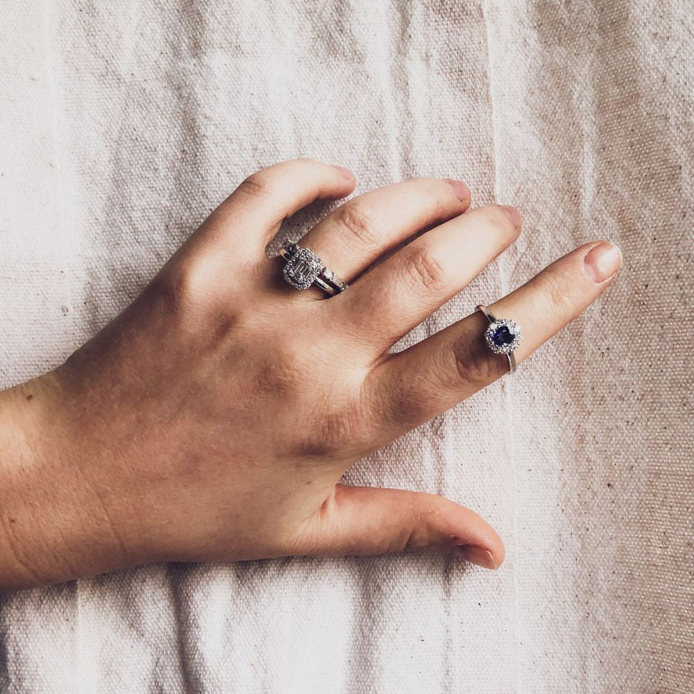 Een hand op lichte stof draagt een Elegante Witgouden Ring met Briljant en Baguette Diamanten (0,54 crt) van Excellent Jewelry aan de middelvinger, in combinatie met een zilveren ring met een donkerblauwe steen aan de ringvinger.