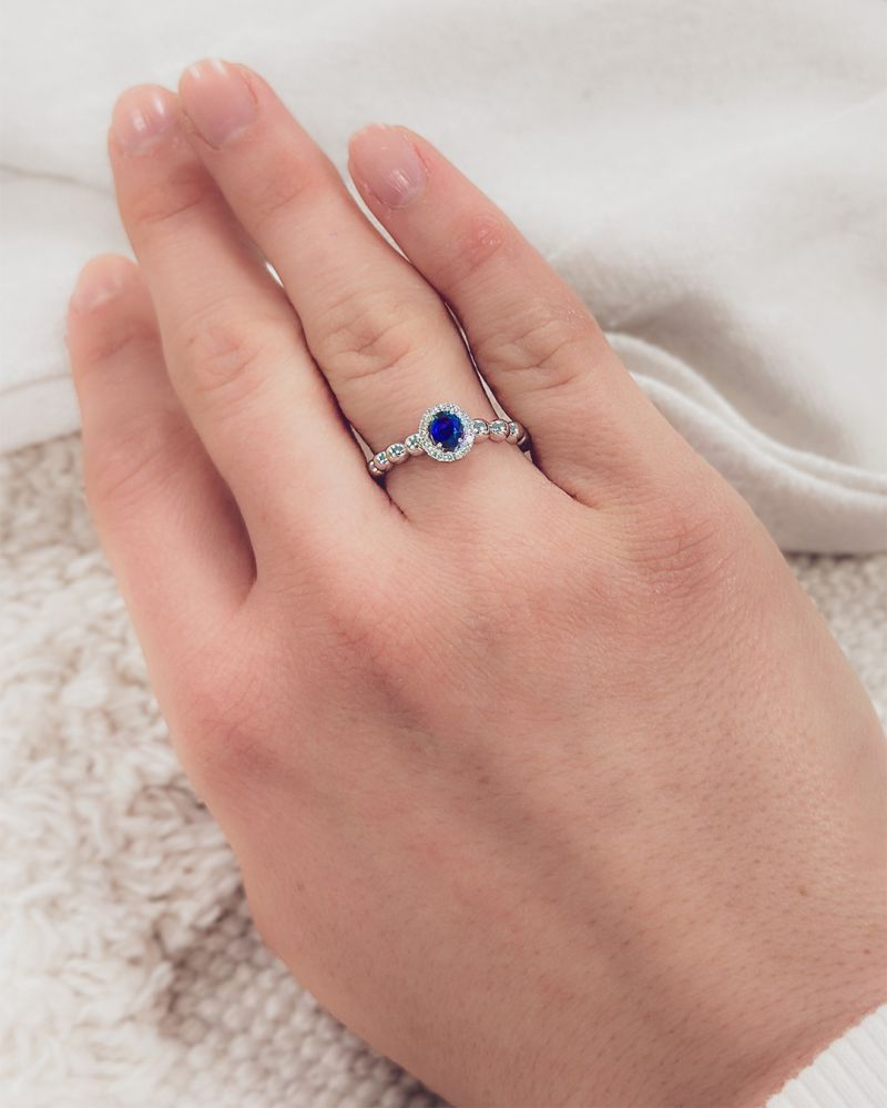 Een goed gemanicuurde hand showt de 14 Karaat Witgouden Rijring met Blauwe Saffier en 0,14 Caraat Diamanten van Excellent Jewelry op een gestructureerd wit oppervlak.