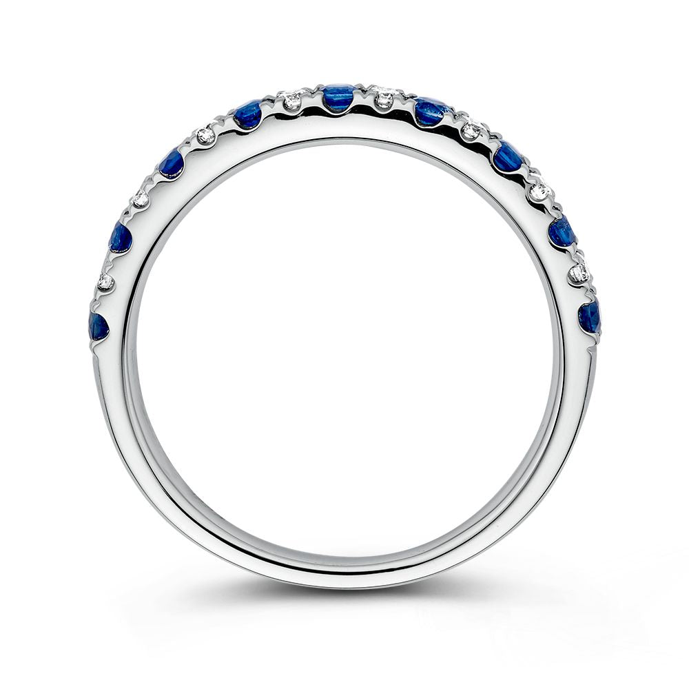 De 14 karaat witgouden rijring van Excellent Jewelry heeft afwisselende blauwe saffier en 0,14 ct diamanten langs de bovenkant van de band, getoond van opzij tegen een witte achtergrond.