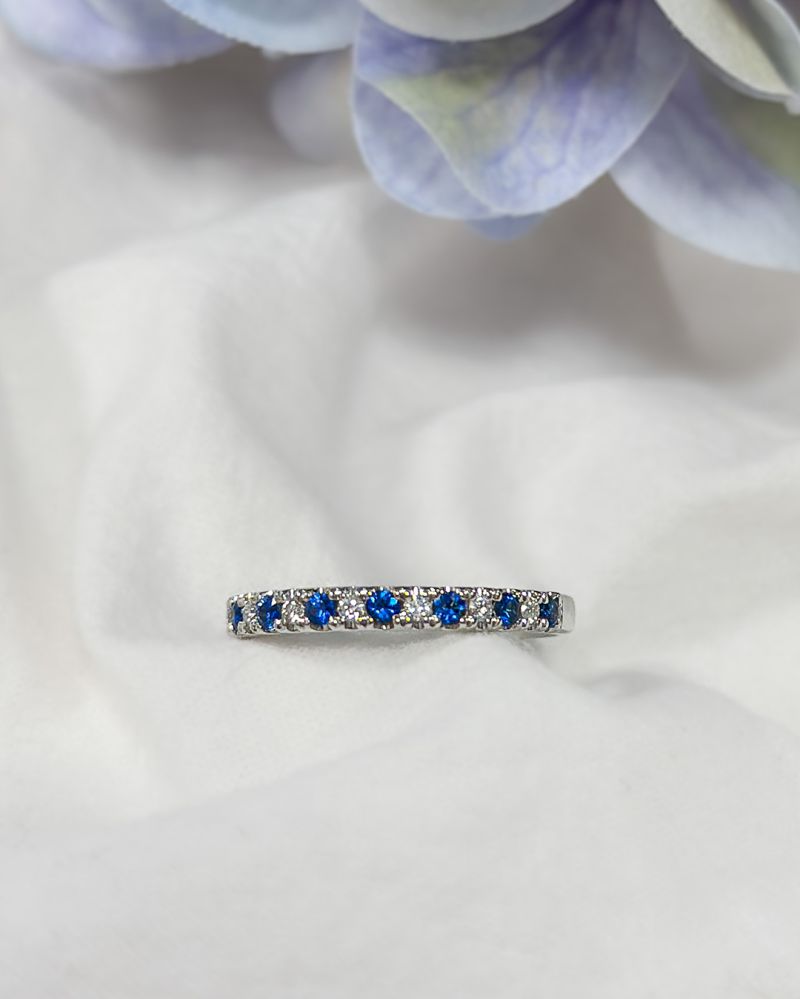 De 14 karaat witgouden rijring met blauwe saffier en 0,14 ct diamanten van Excellent Jewelry schittert op een zacht wit kleed, omgeven door paarse en groene bloemblaadjes in de vervaagde achtergrond.