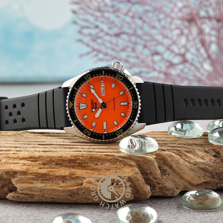 Een Seiko 5 Sports heren horloge SRPL89K1 van Seiko, met een oranje wijzerplaat en zwarte rubberen band, ligt op drijfhout tussen glazen steentjes. De achtergrond toont zacht vervaagde blauwe en roze koraalvormen.