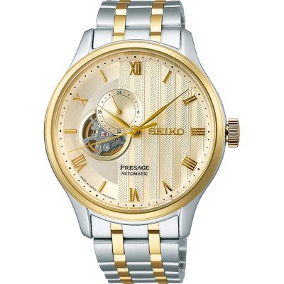 Het Seiko Presage heren horloge SSA464J1 heeft een tweekleurige band van zilver en goud, gouden accenten, een open wijzerplaat waarop het automatische uurwerk te zien is, Romeinse cijfers en structuurdetails.