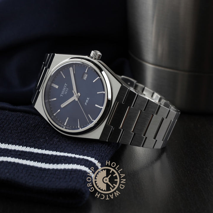 Een Tissot PRX polshorloge van Tissot, met een donkerblauwe wijzerplaat en roestvrijstalen band, rust op marinegestreepte stof. Op de achtergrond staat een metalen tuimelaar met aan de voorkant het logo van Holland Watch Group.