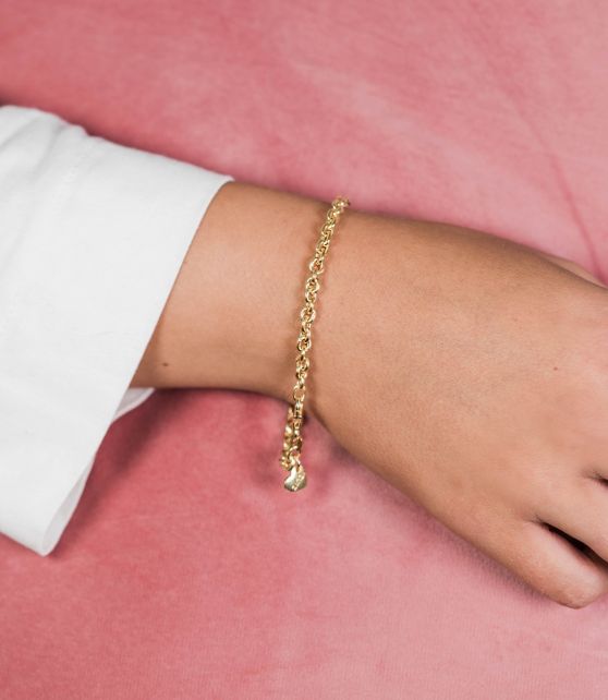 Een close-up van de hand van een persoon op een roze oppervlak, met daarop de Zinzi Armband Gold Plated Zia1153 G. Deze delicate gouden armband vormt een elegante aanvulling op hun witte shirt met lange mouwen.